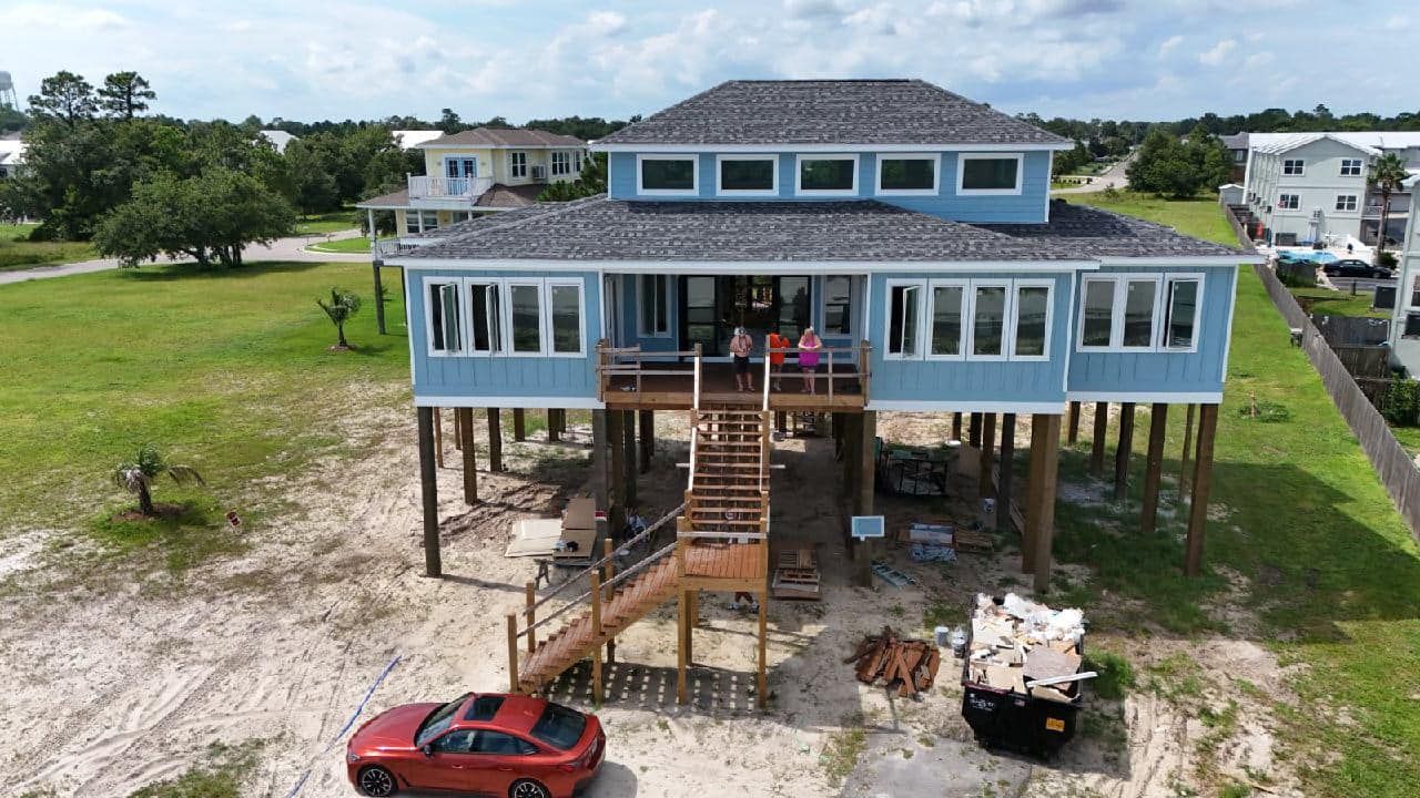 Blue coastal home on stilts, with a red car parked in front. People are visible on the deck.