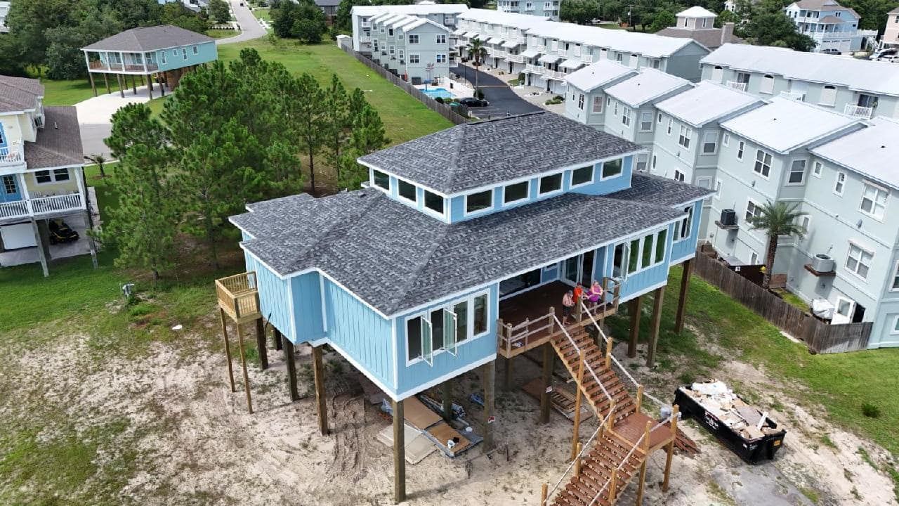 Elevated blue beach house with a multi-tiered roof and wooden stairs, set on sandy ground. Two people are on the deck.