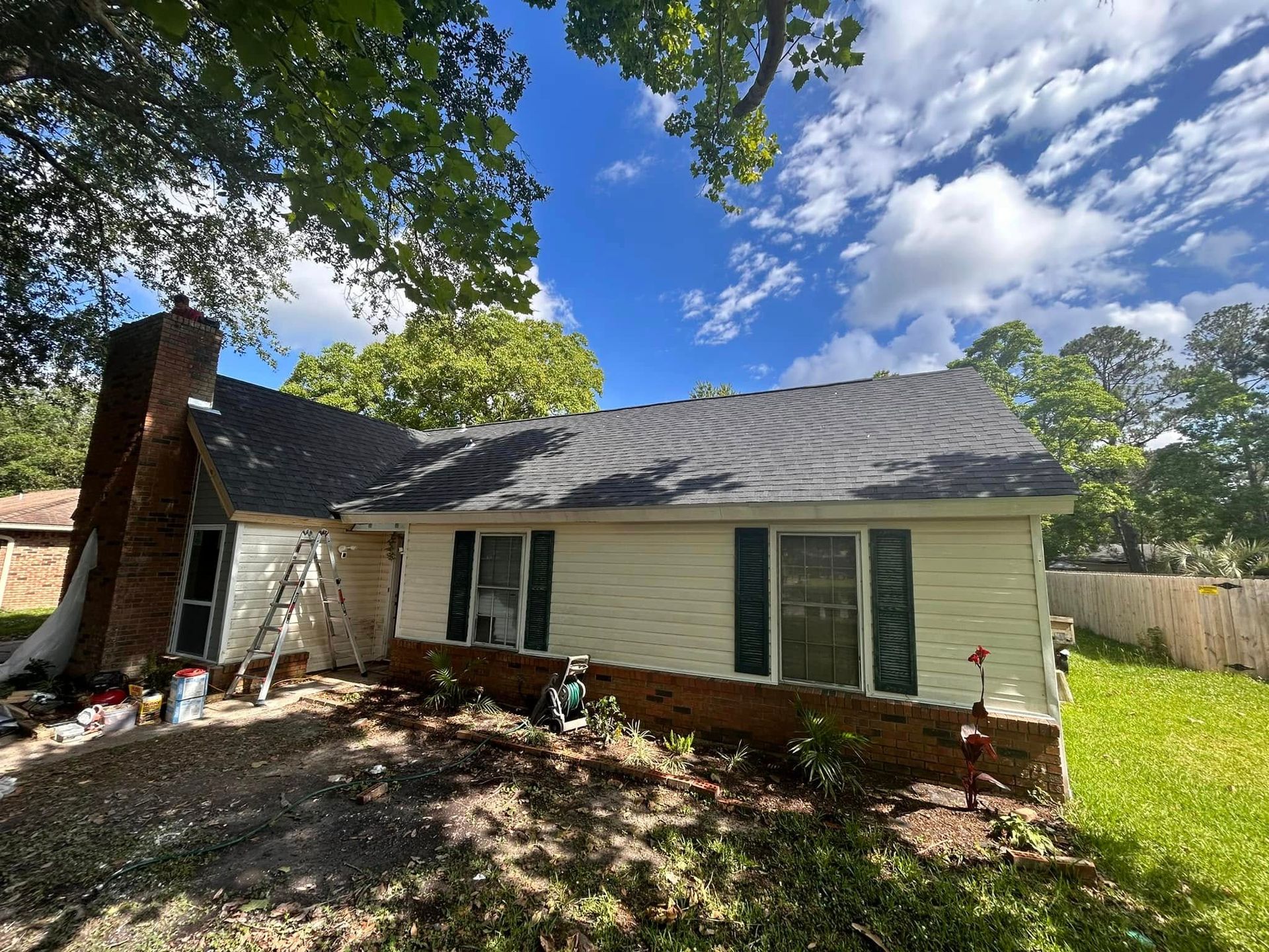 Beige house with a dark roof, brick chimney, and green shutters. A ladder leans against the side, with trees and a blue sky visible.