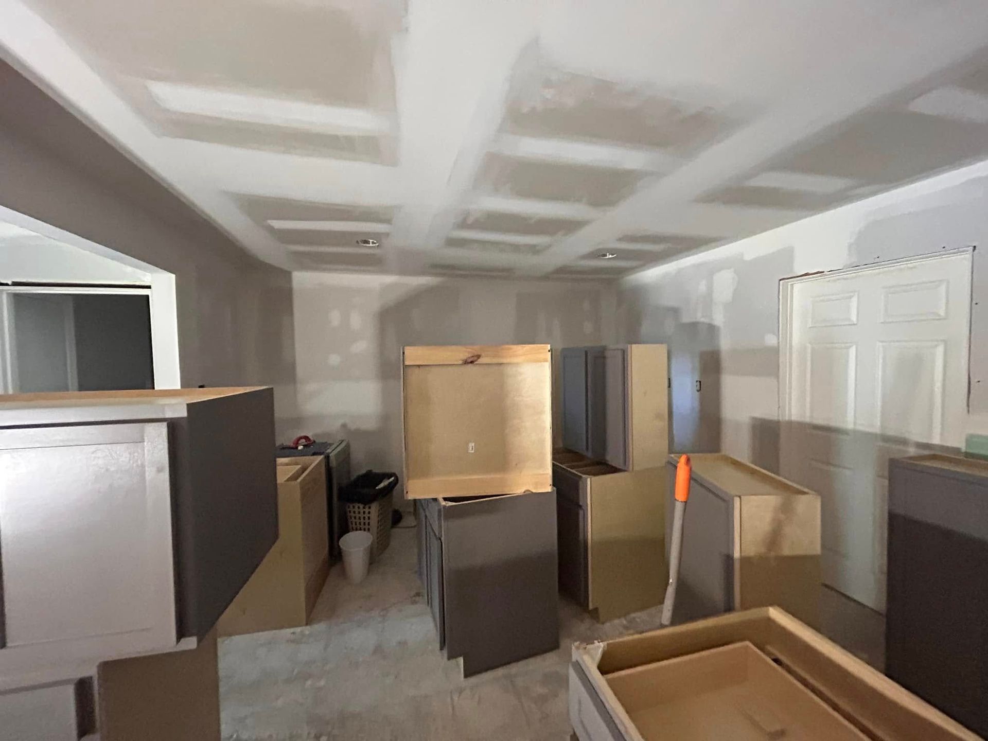Kitchen under construction with drywall, gray cabinets, and uninstalled cabinets scattered around.