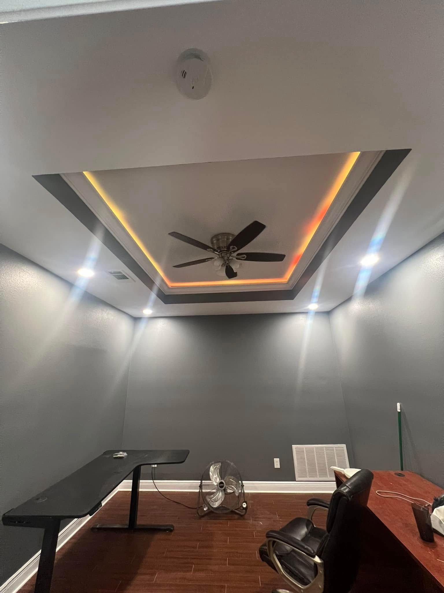 Room with dark gray walls, a dark ceiling fan, and a recessed ceiling with colorful LED lighting. A long black desk is on the left.