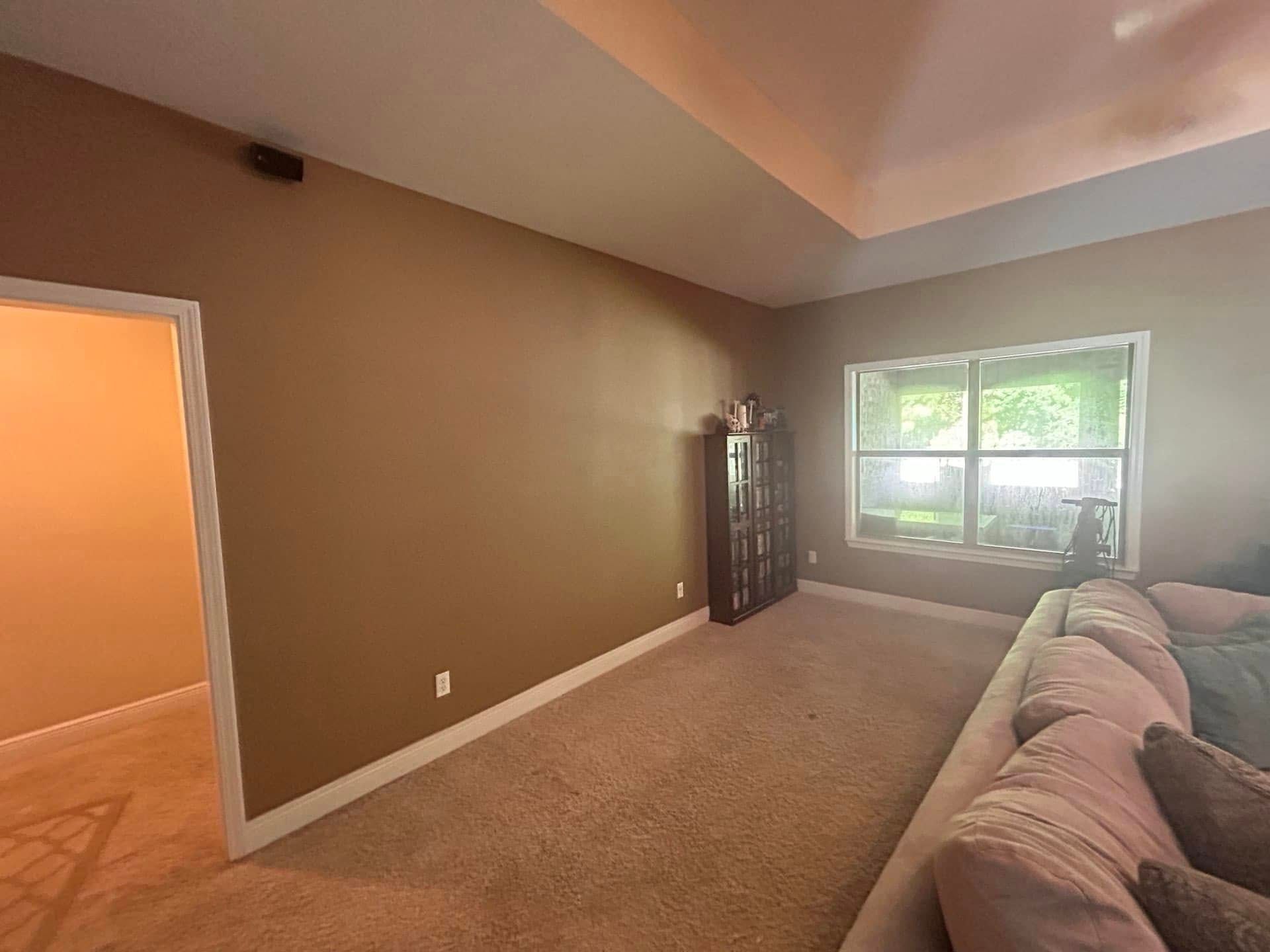 A living room with a brown wall, carpet, and a built-in cabinet.  A window with a view of greenery is on the right.