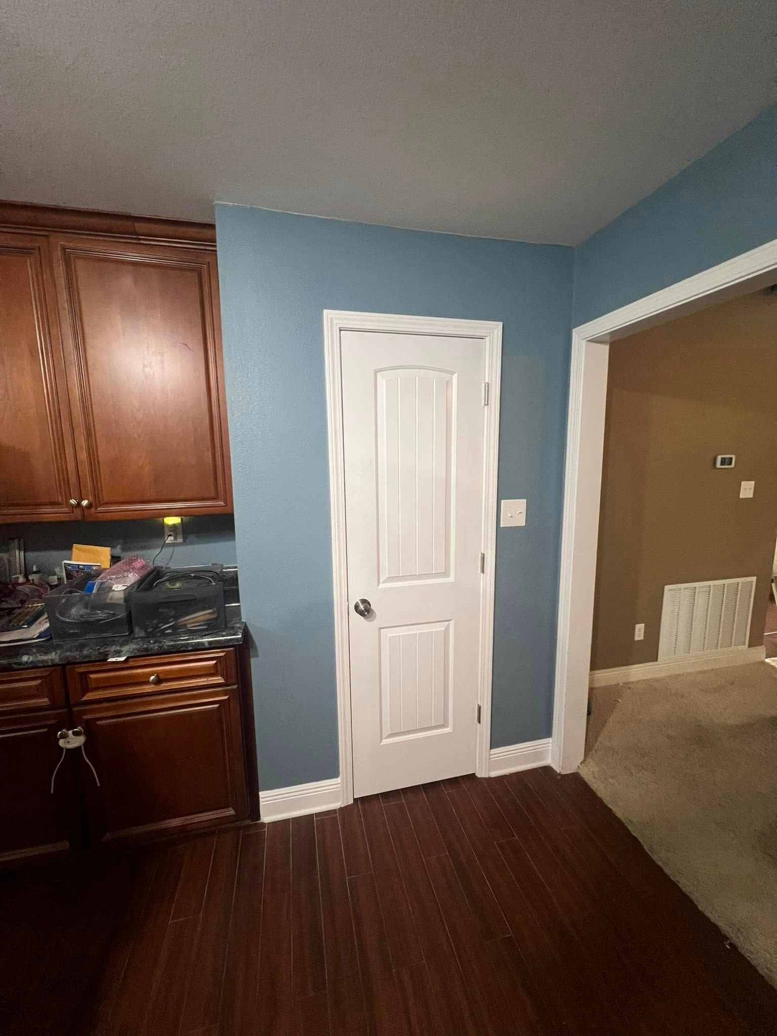 A kitchen with a wooden floor, dark brown cabinets, and a white door surrounded by blue walls.  An adjacent room is visible to the right.