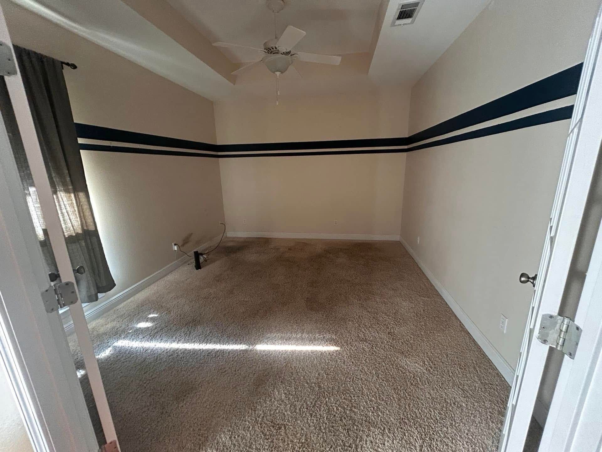 An empty beige-walled room with dark blue stripes. The room has a ceiling fan, brown carpet, and a partially visible window with a curtain.