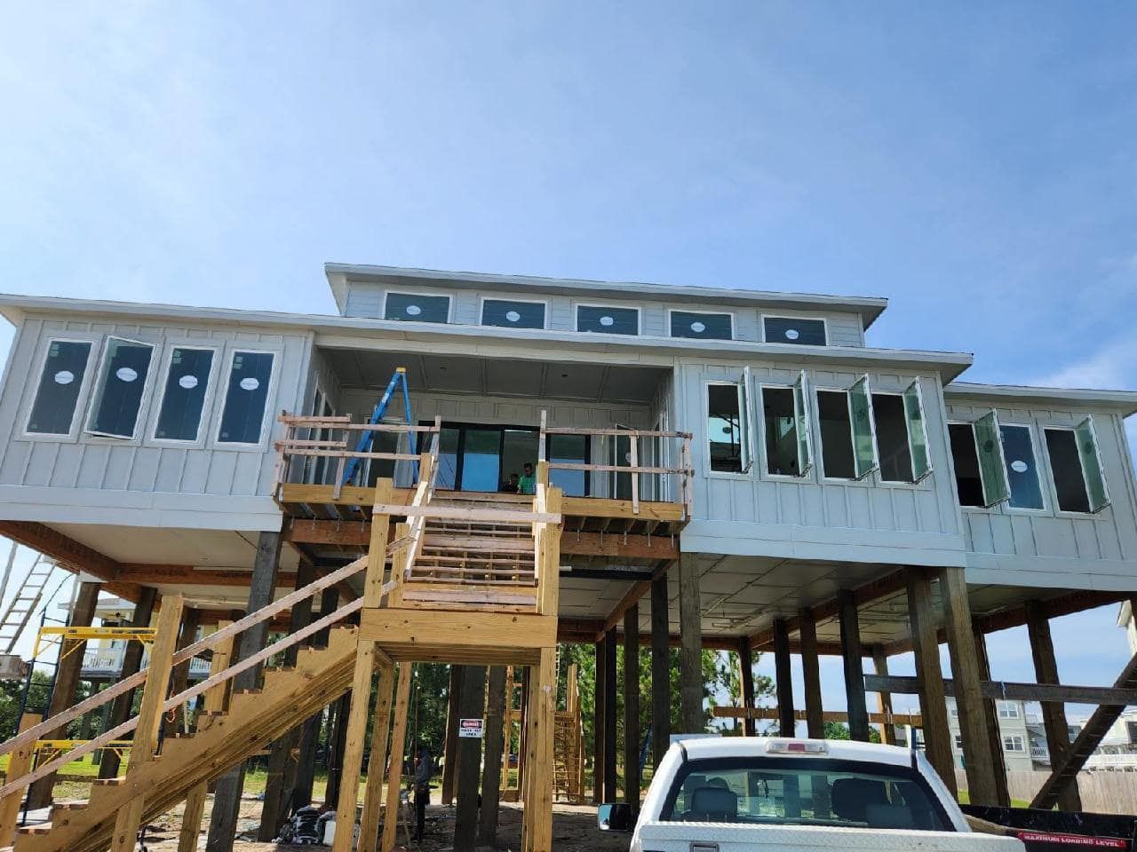 A light gray, stilt house under construction with a wooden deck and staircase. A white pickup truck is parked in front on a sunny day.