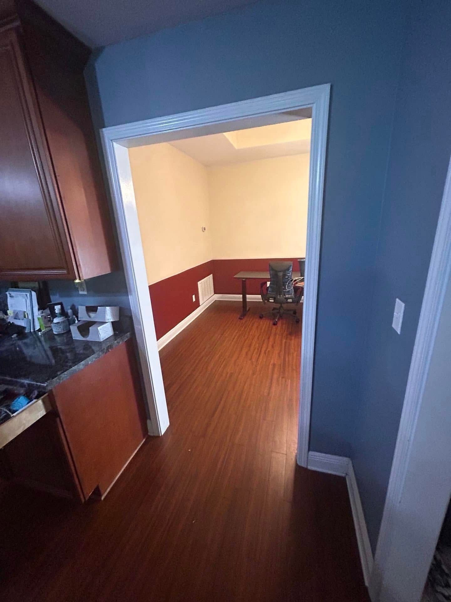 View from a kitchen doorway into a room with red and yellow walls, a desk, and a chair. Brown cabinets and blue wall are in the foreground.