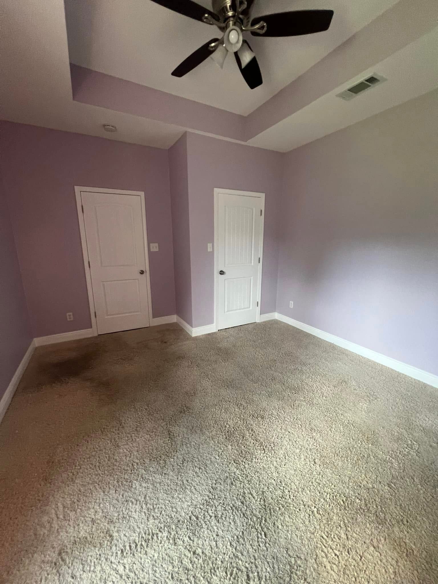A bedroom with purple walls, two white doors, and beige carpeting. A black ceiling fan is mounted on the white ceiling.