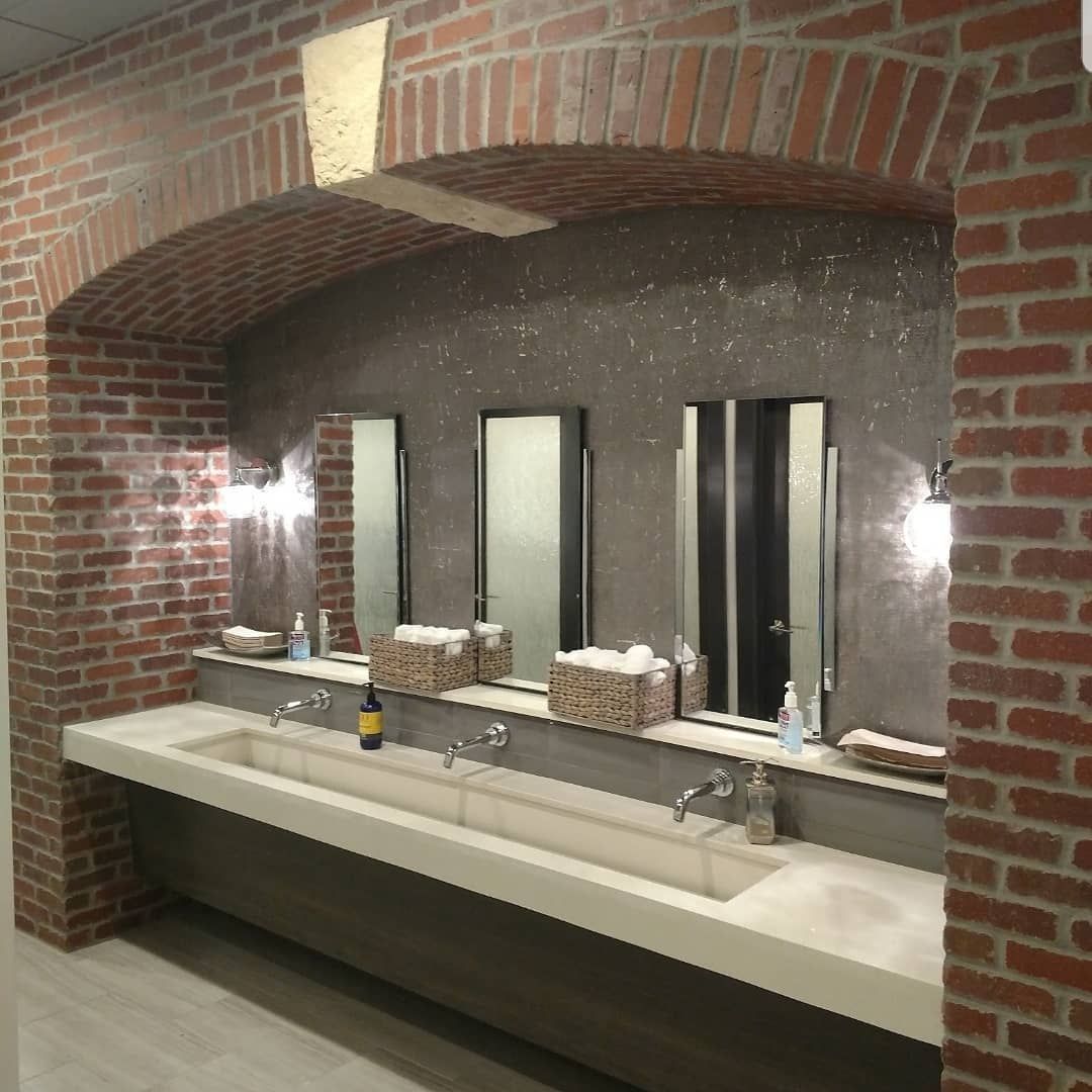 Brick-walled restroom with long trough sinks, mirrors, and an arched brick doorway.