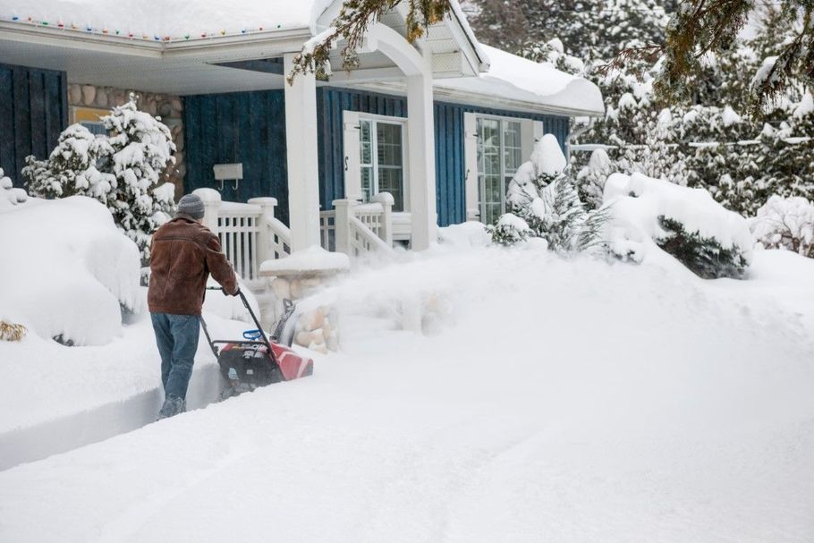 Person clearing deep snow from a driveway outside a blue house