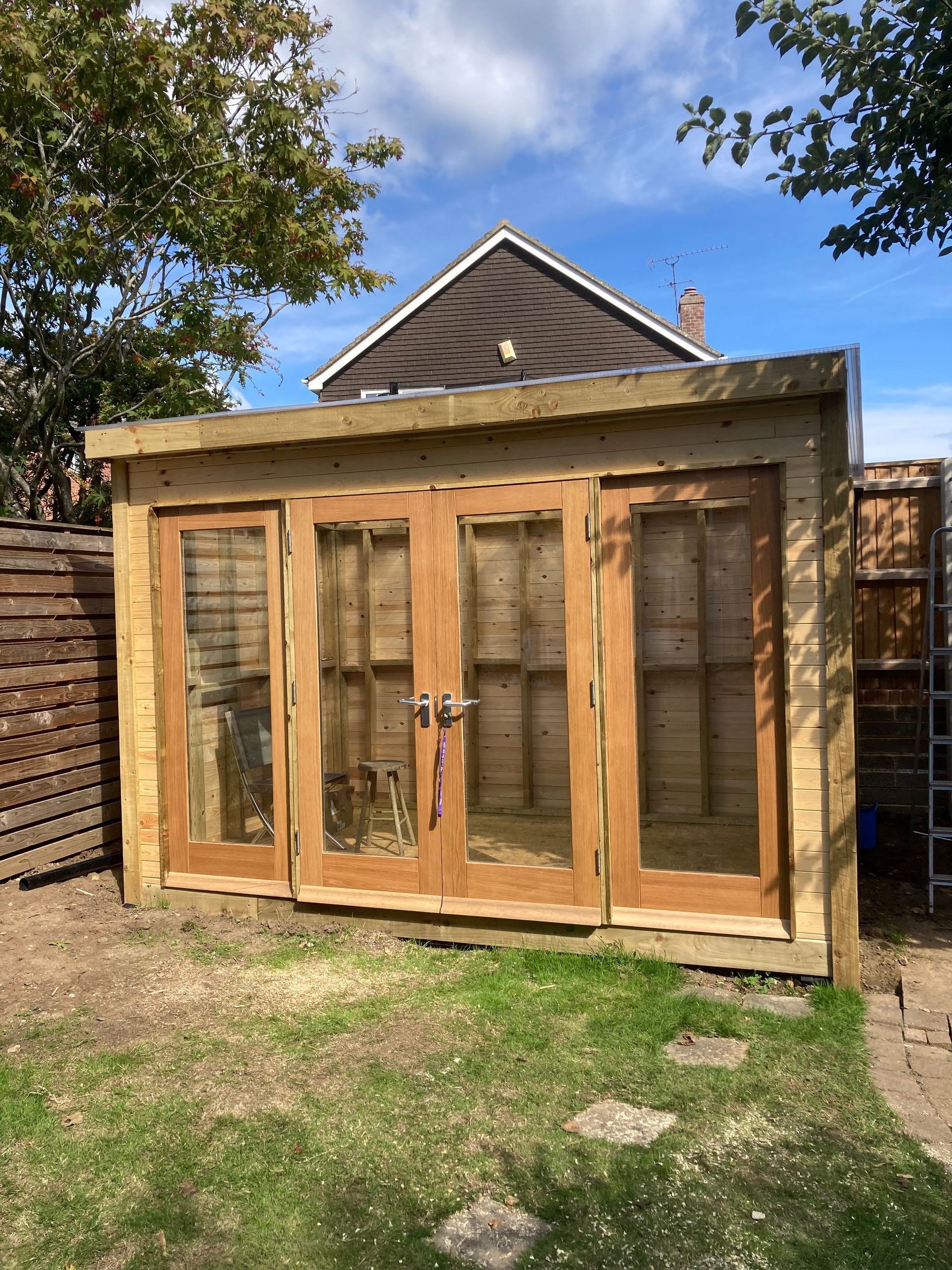 Wooden garden shed with glass double doors and side windows in a grassy backyard.