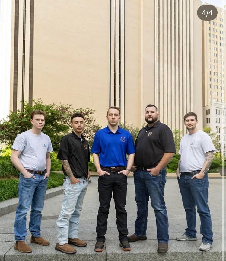 Five men in casual attire pose outside a tall building. Some have tattoos.