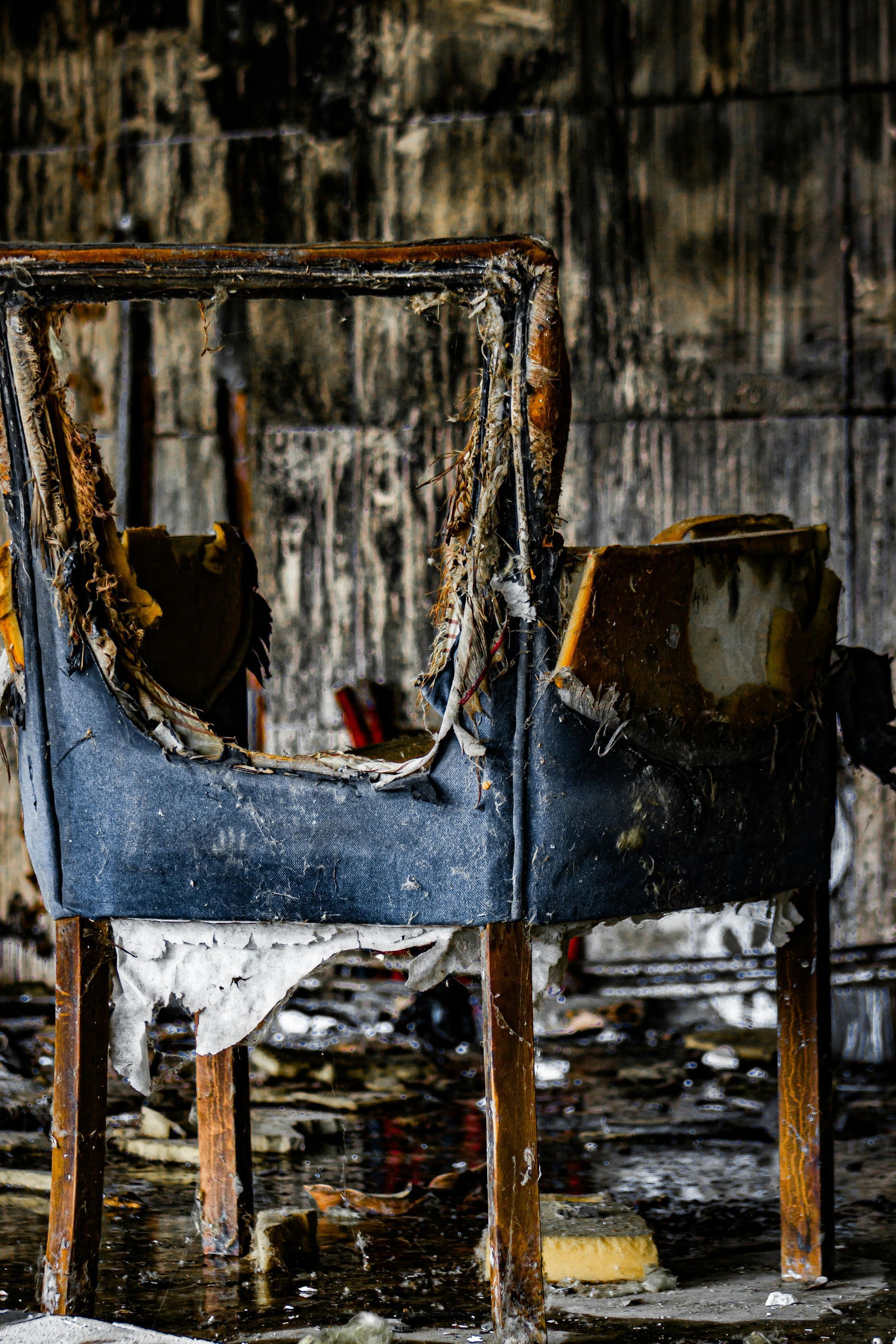 Burnt, damaged chair frame with blue upholstery in a fire-ravaged room; wood paneling visible.