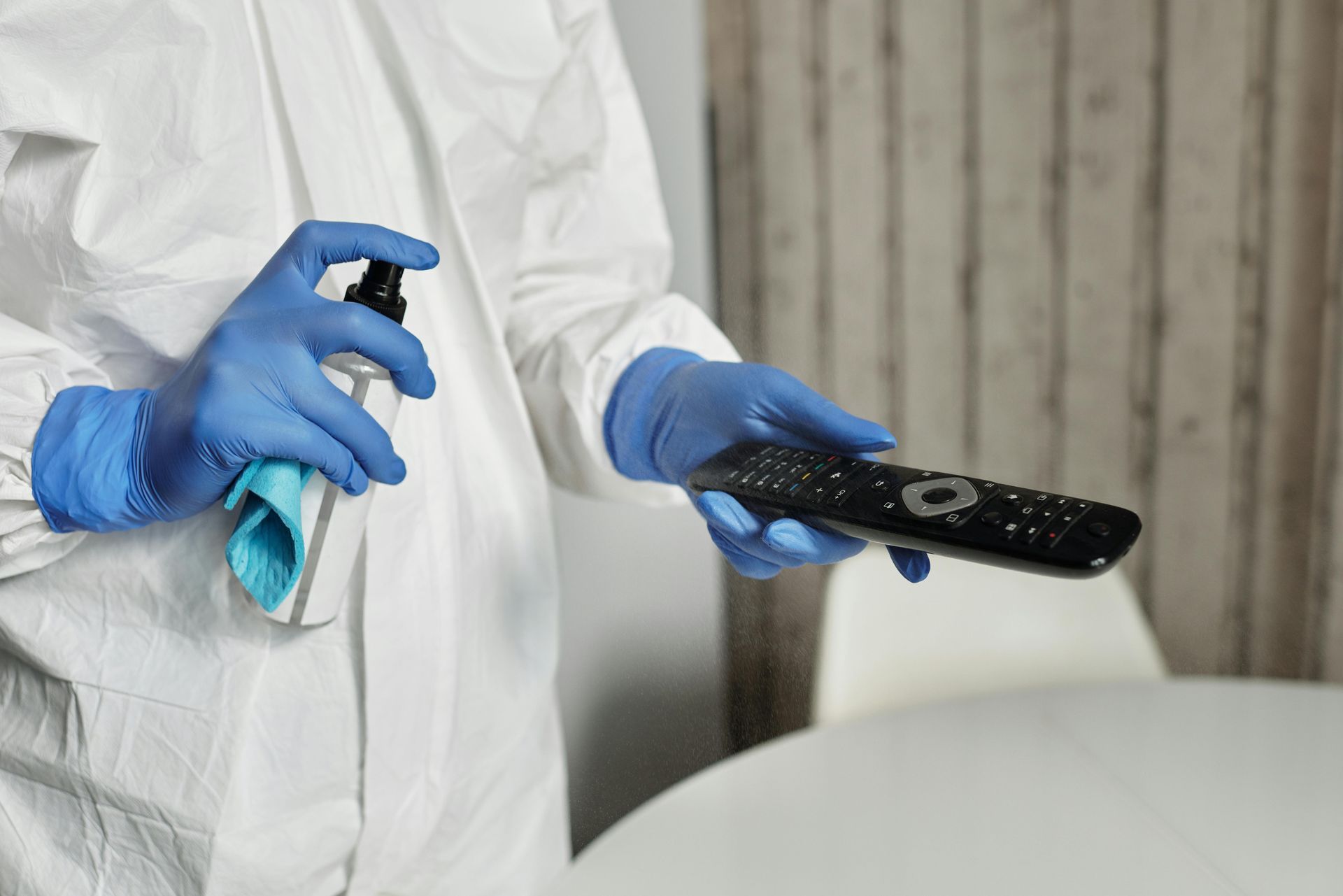 Person in protective suit spraying a remote control with a blue gloved hand in an indoor setting.