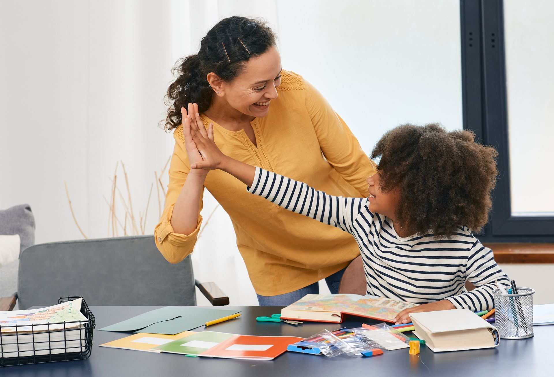 Woman and child high-fiving at a table with school supplies; both smiling in a brightly lit room.
