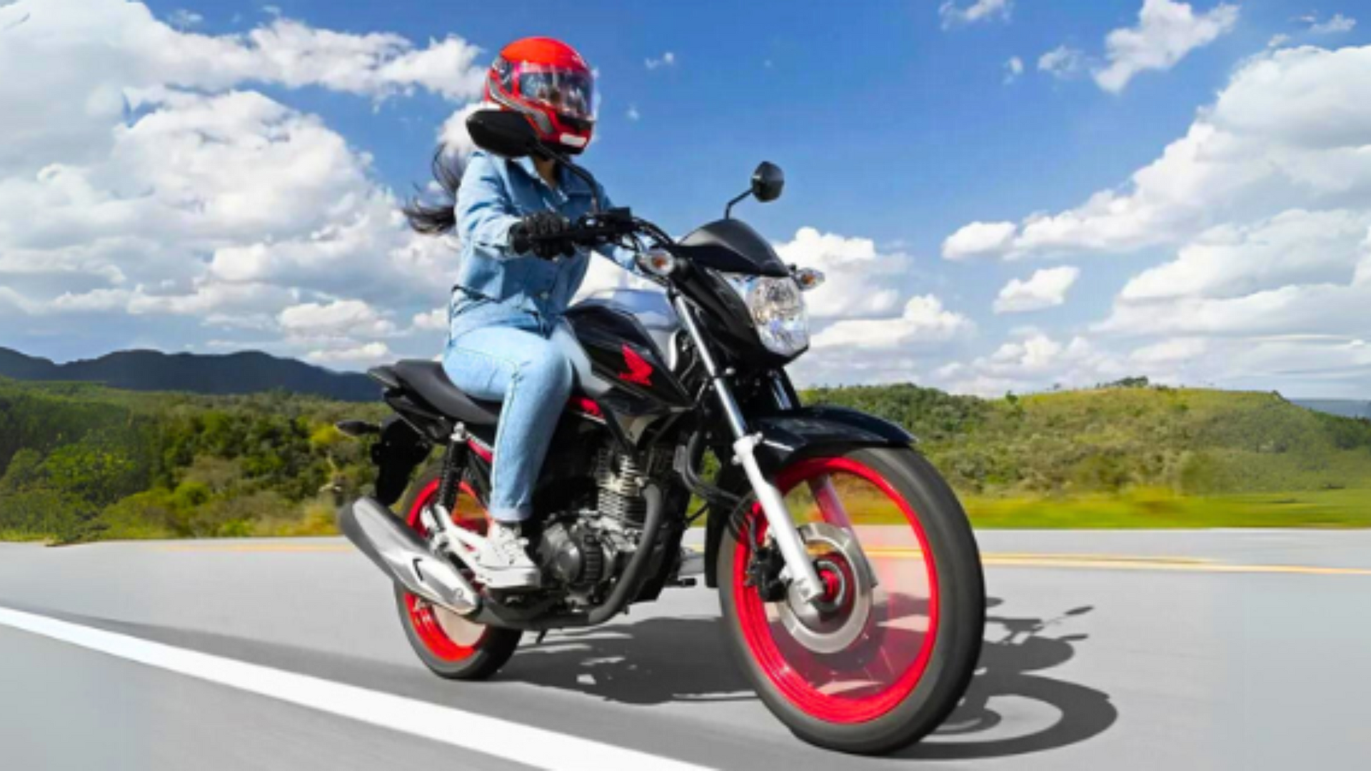 Mulher pilotando uma motocicleta preta em uma estrada ensolarada, usando um capacete vermelho e uma jaqueta jeans azul.