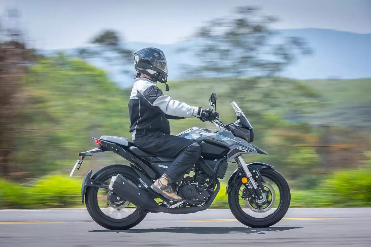 Motociclista de preto pilotando em uma estrada, com árvores verdes desfocadas ao fundo.