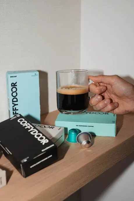 A hand holds a glass of espresso over a shelf with coffee capsule boxes. The boxes feature Coffybox™ branding.
