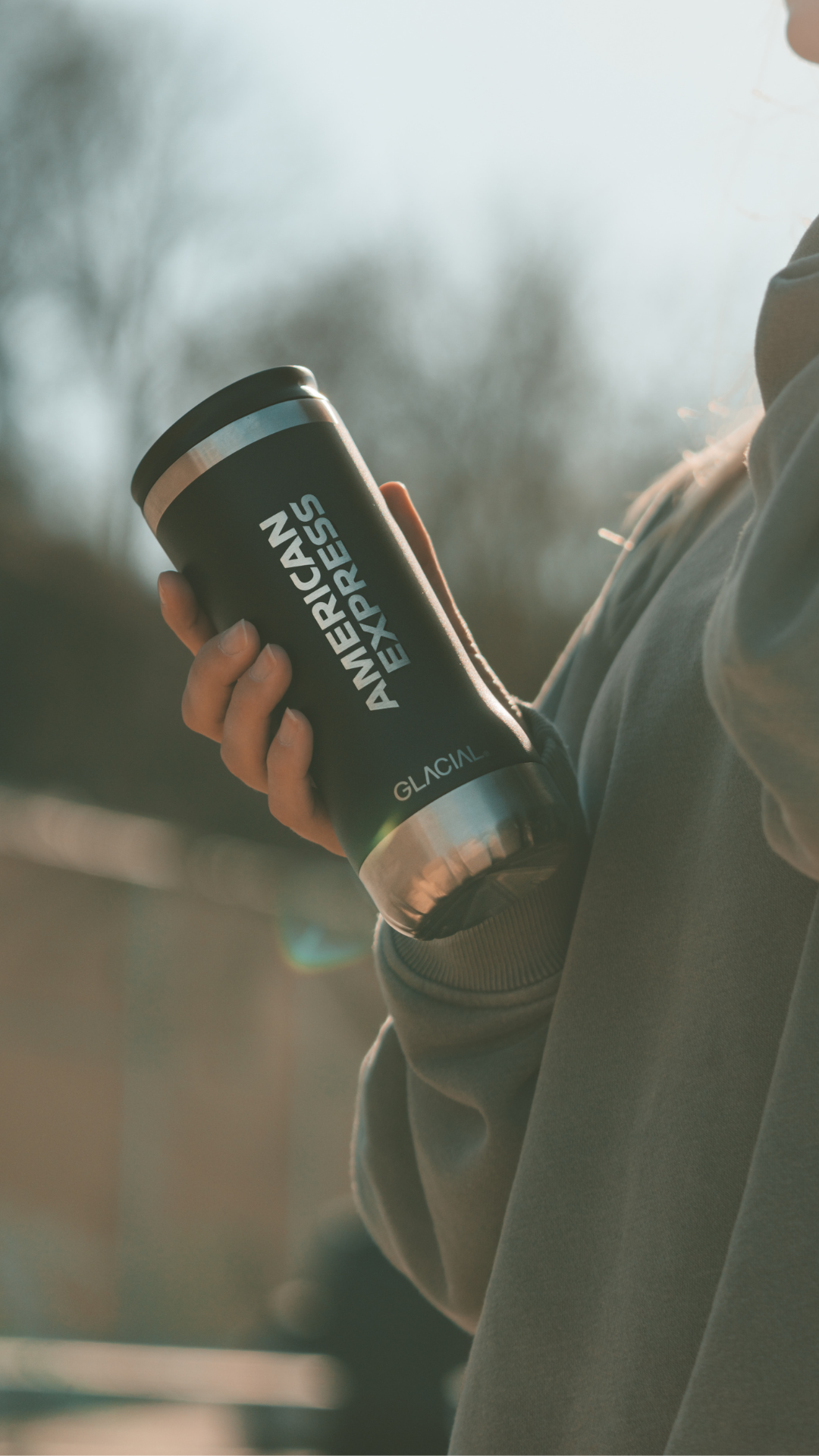 Person outside holding a black coffee tumbler with American Express™ and Glacial™ branding.