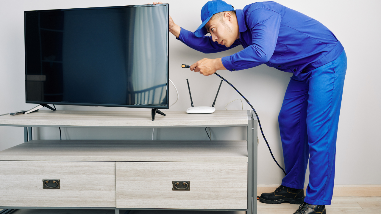 A person in blue overalls plugs a power cord into a Wi-Fi router on a TV stand, near a TV.