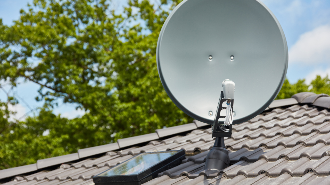 Satellite dish mounted on a tiled rooftop, positioned against a backdrop of trees and a blue sky.