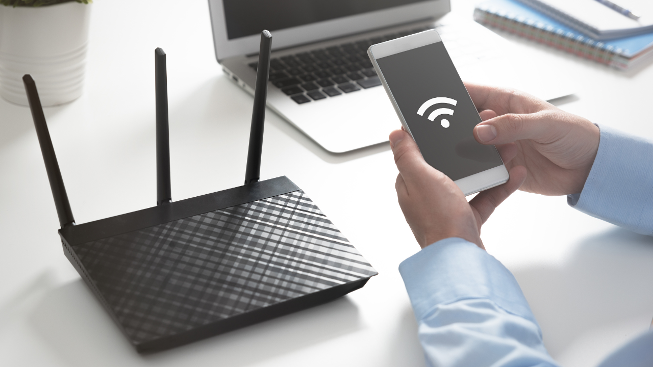 Black router next to a person holding a phone displaying a Wi-Fi signal, near a laptop and notepad.