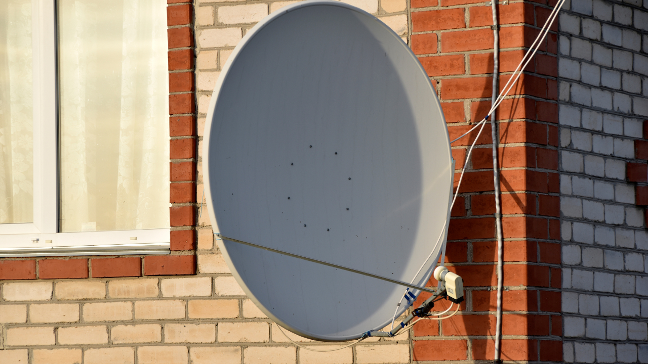 A satellite dish, gray, mounted on a brick wall next to a window.