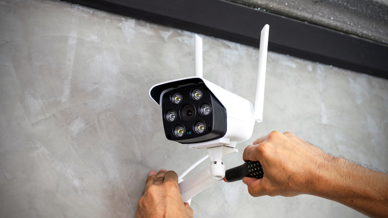 A person installing a white security camera with antennas on a concrete wall.