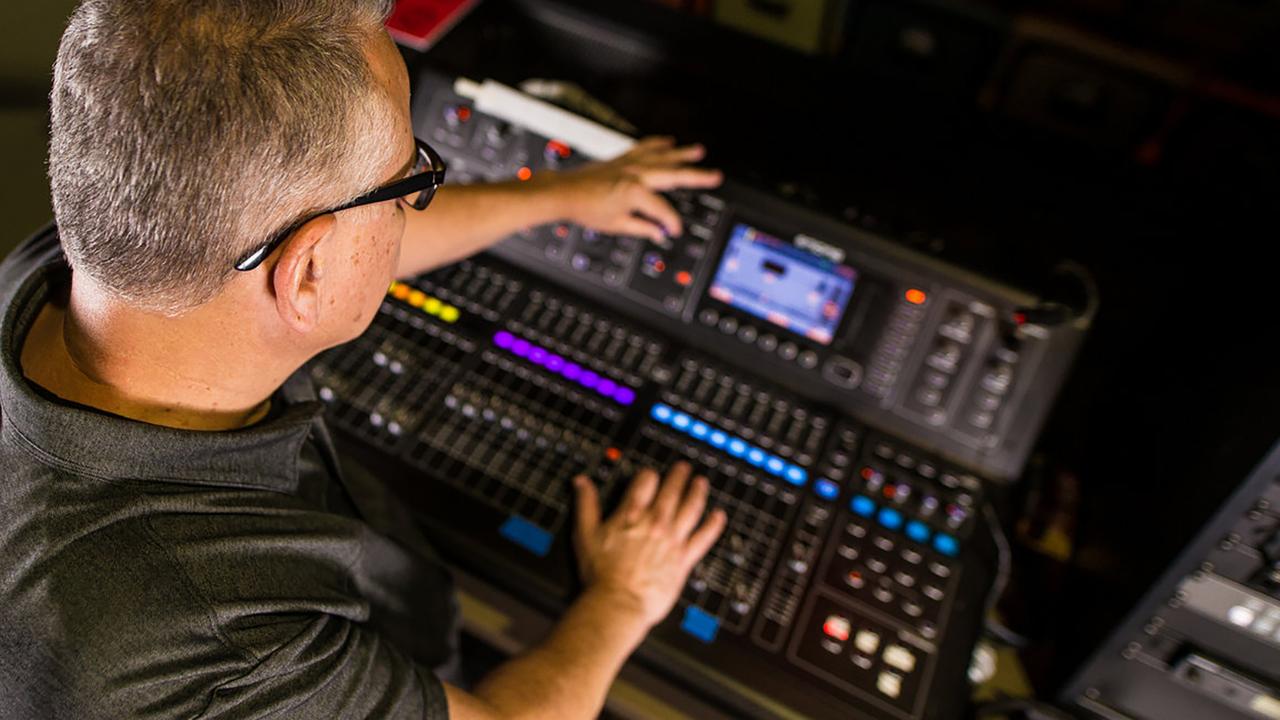 Man operating a sound mixing console with multiple faders and a screen, in a dim environment.