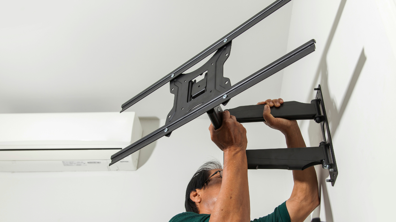 Person installing a black articulating TV mount on a white wall; air conditioner visible.