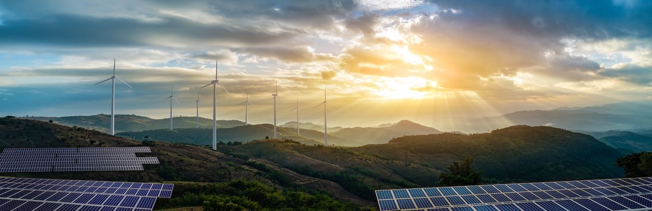 Solar panels in foreground, wind turbines on a mountain ridge, sun shining through clouds.