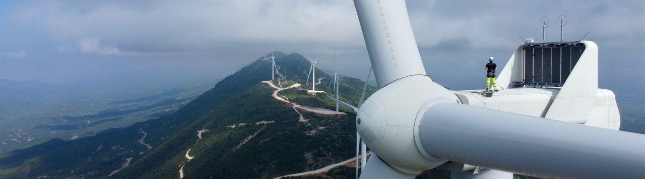Person in safety gear on top of a wind turbine, overlooking a rural landscape under a cloudy sky.