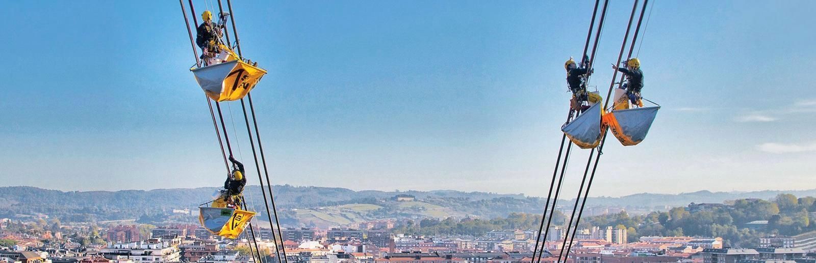 Cable stay workers in suspension on ropes with buckets underneath, against a clear blue sky, overlooking a city.