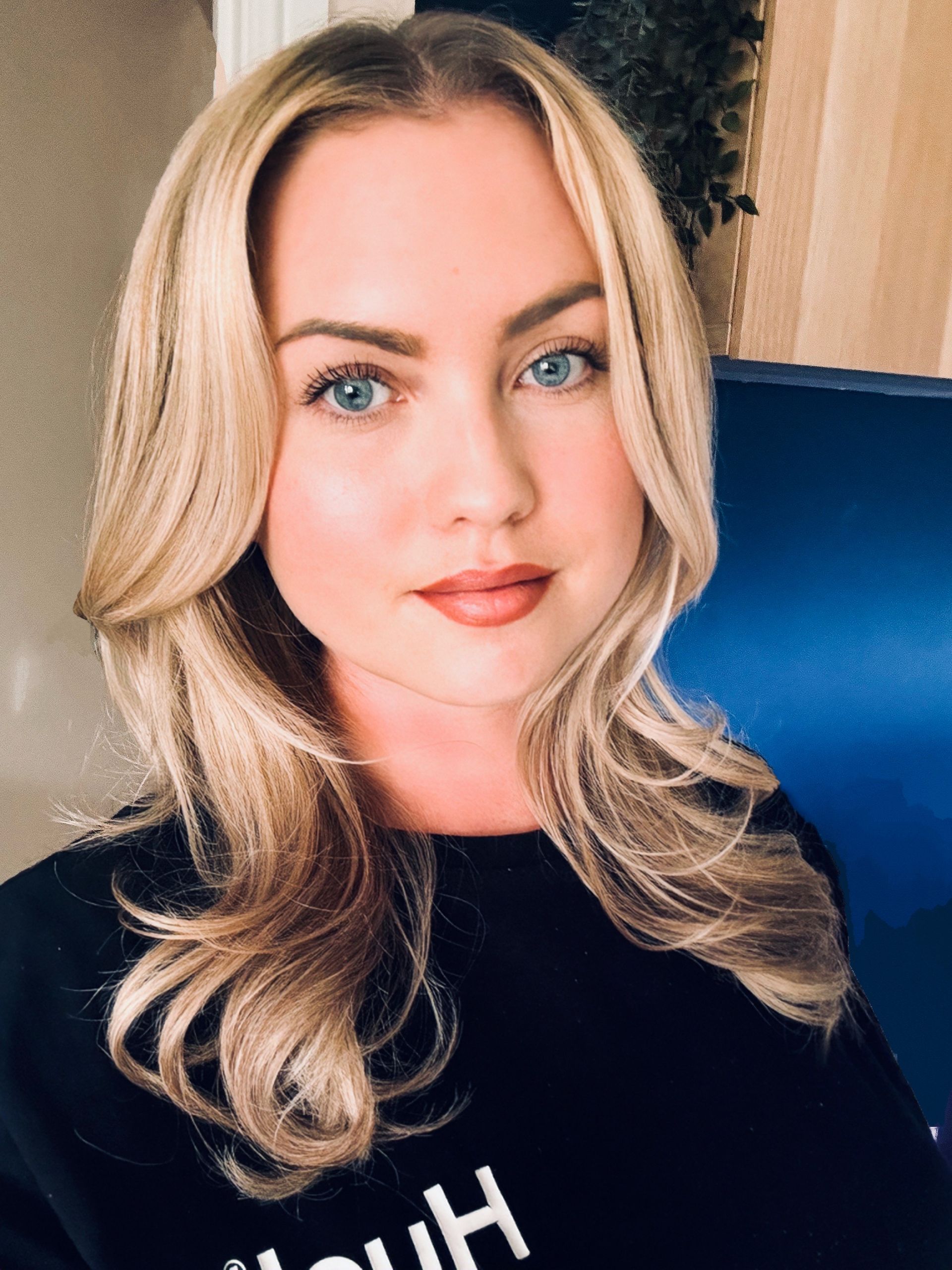 Sophie is Blonde with blue eyes and light makeup, smiling slightly. Wearing a black top. Blue background.