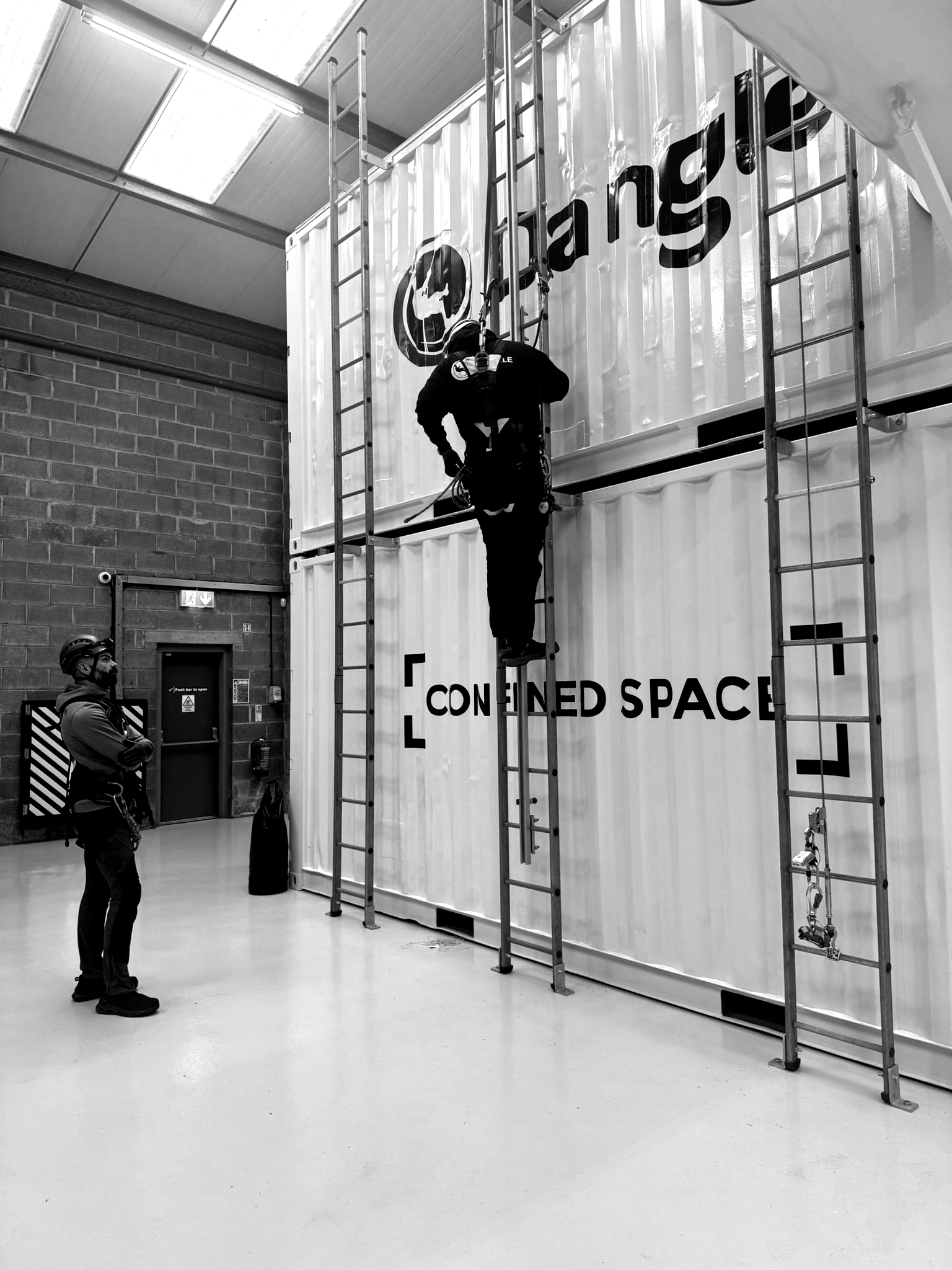 A person in safety gear climbs a ladder against a training container at Dangle Academy, observed by another person.