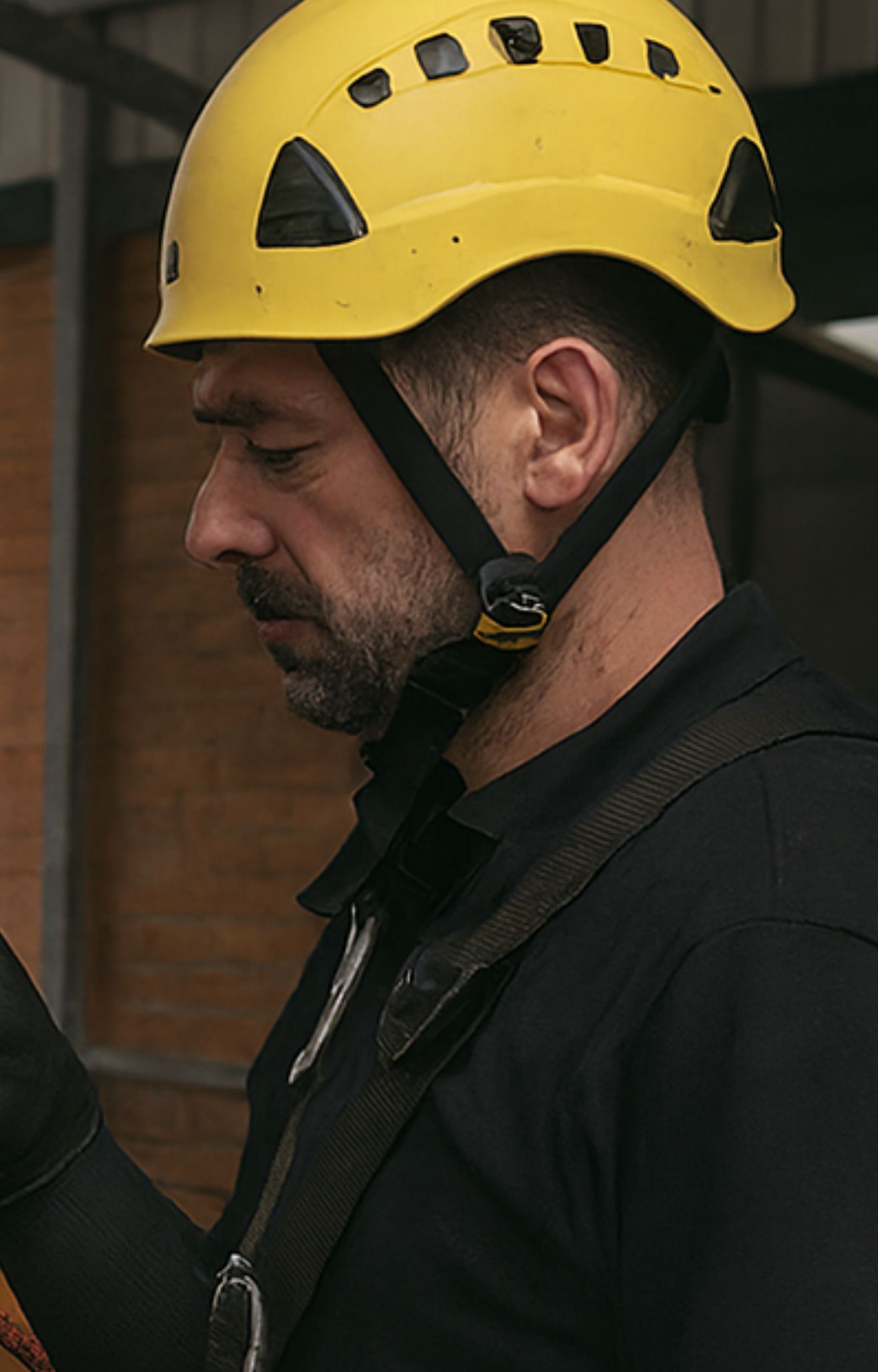 Man in yellow hard hat and black shirt, focused, indoors.