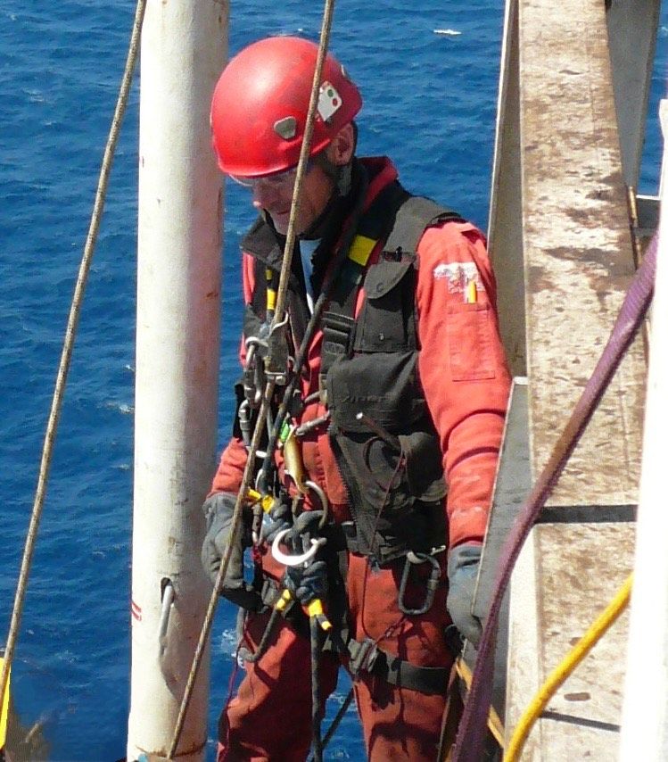 Bruce in red safety gear on a structure over blue water. He's wearing a helmet and harness, likely for rescue.