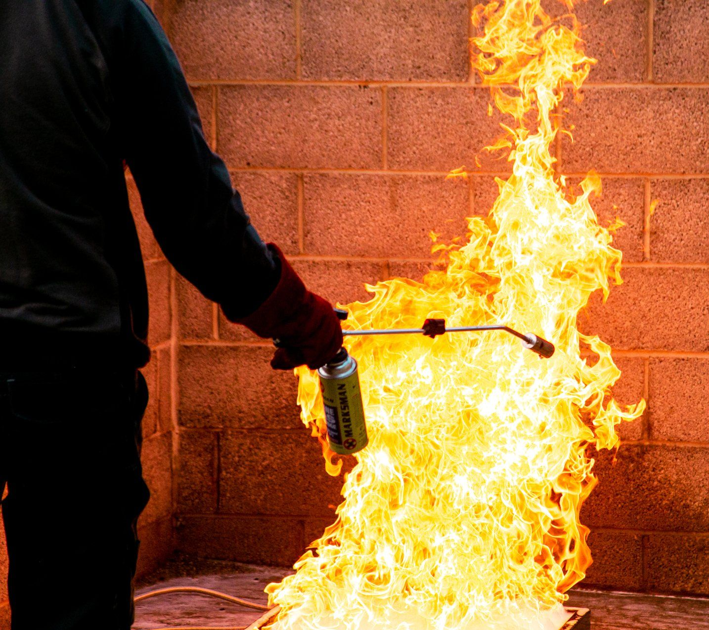 Person in gloves using a torch, large flames against a brick wall.