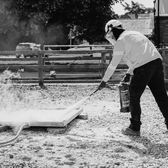 Worker in protective gear using a fire extinguisher on concrete outdoors.