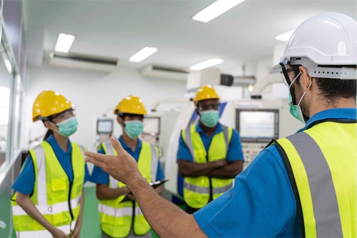Group of workers in safety gear and face masks, one speaking, in a factory setting.