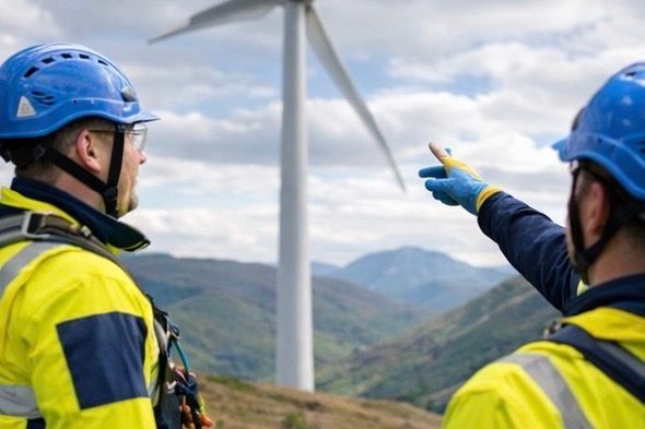 Two workers in safety gear point towards a wind turbine on a mountain, blue sky.