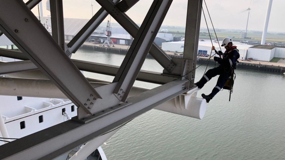 Worker rappelling from a metal structure over water. He wears a harness, helmet, and work clothes. Industrial setting.