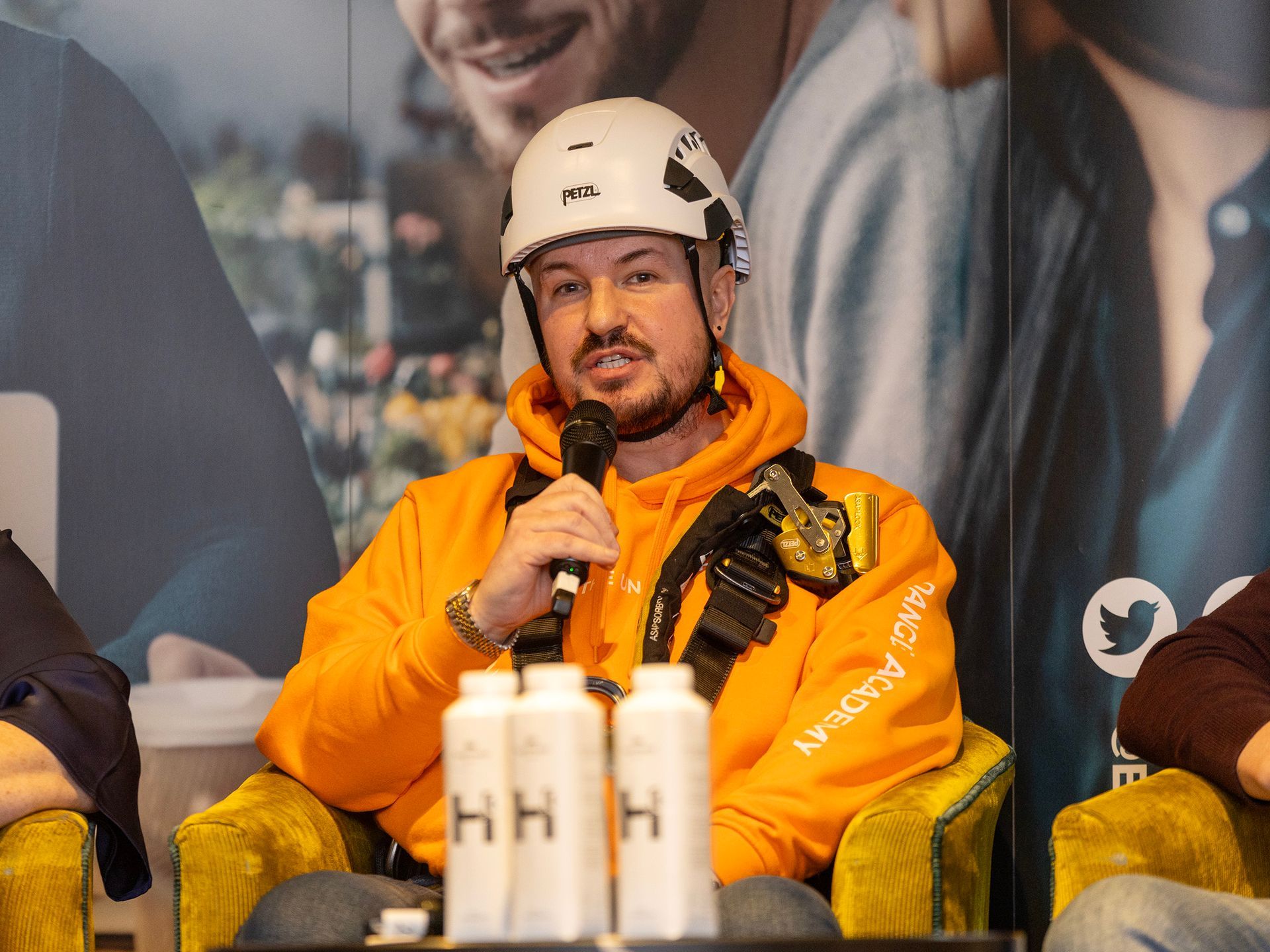 Simon in a orange Dangle hoodie and white helmet speaks into a microphone at a event in Belfast, sitting at a table with water bottles.