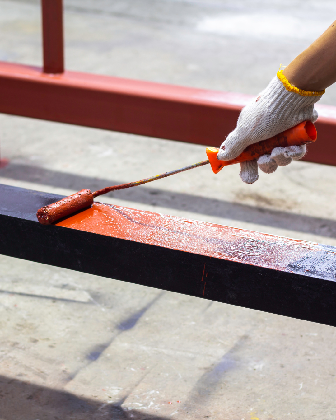 A gloved hand uses a paint roller to apply red paint to a metal beam, indoors.