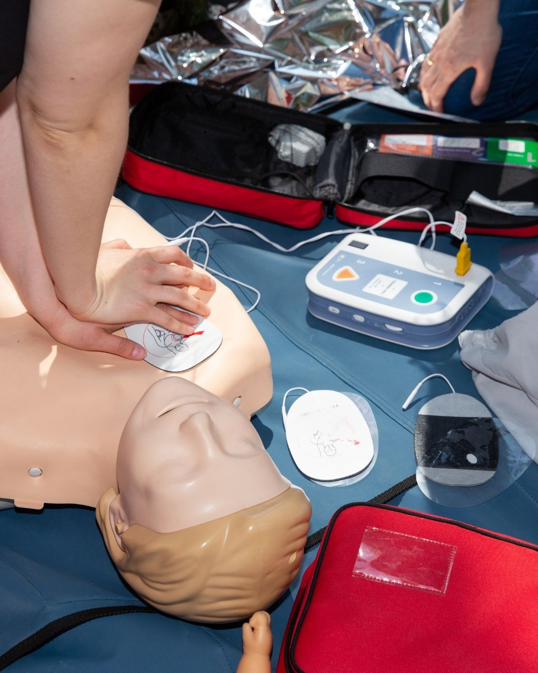 A person performs CPR on a training mannequin next to an automated external defibrillator (AED) and medical supply kits.