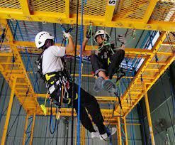 Two people in helmets and harnesses on an indoor adventure course, one climbing, the other seated, yellow framework.