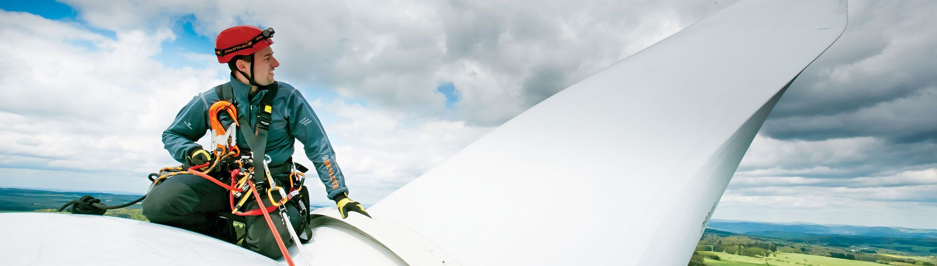 A dangle wind turbine technician in a safety harness on a wind turbine blade, looking out at a cloudy sky.