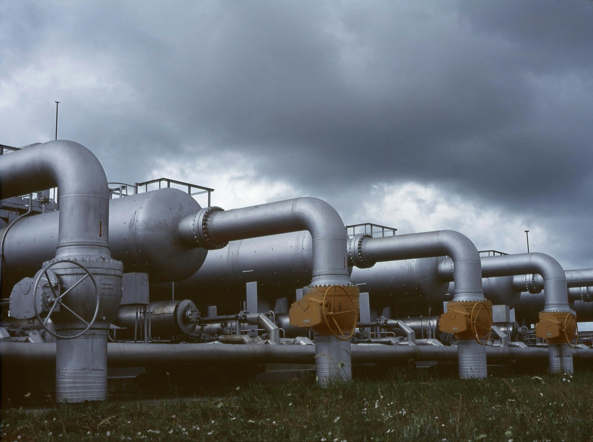 Large metal pipes and tanks against a cloudy sky, in an industrial petrochemical setting.
