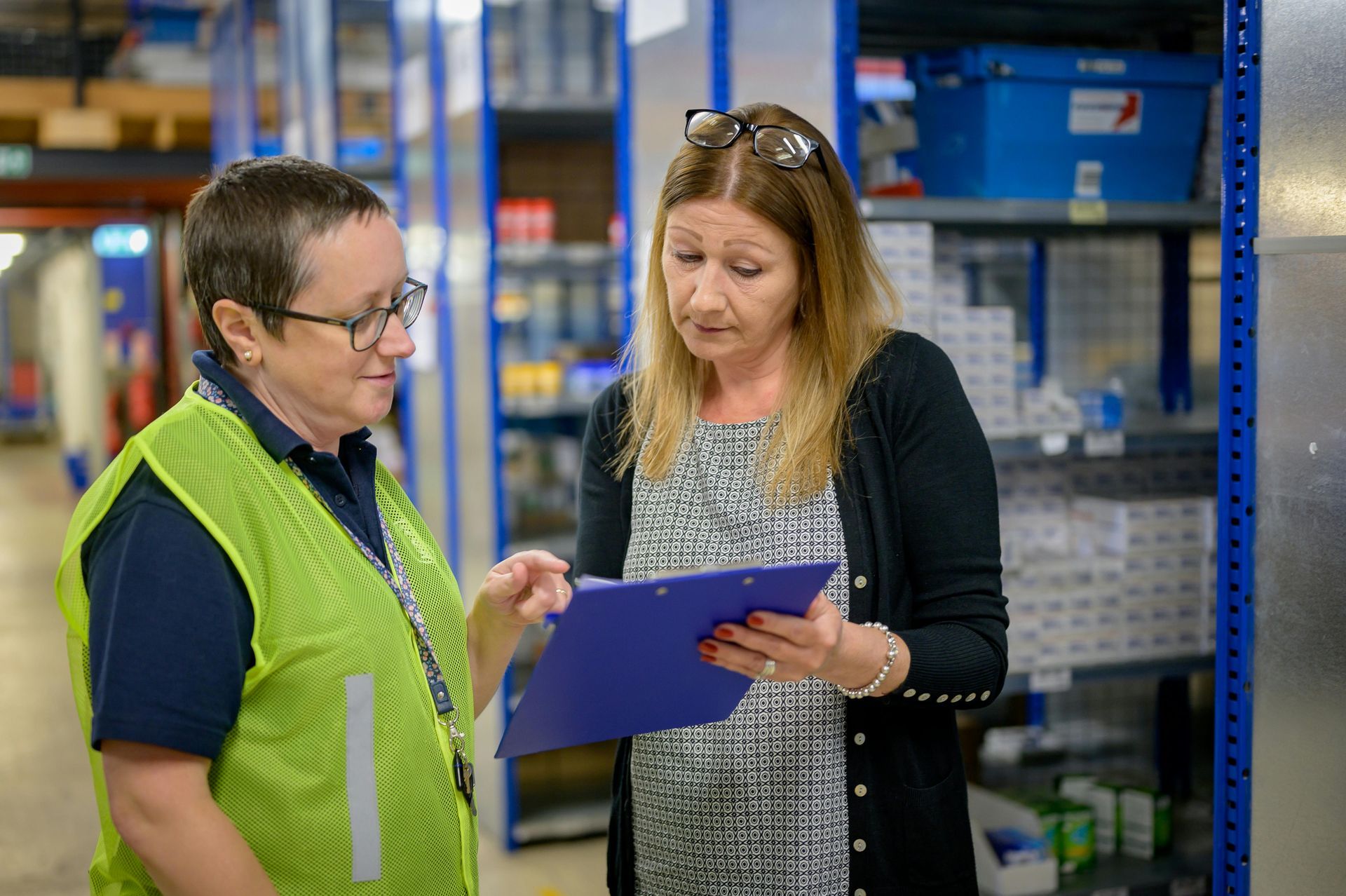 Two women in a warehouse looking at a clipboard. One wears a safety vest. Shelves and equipment are visible.