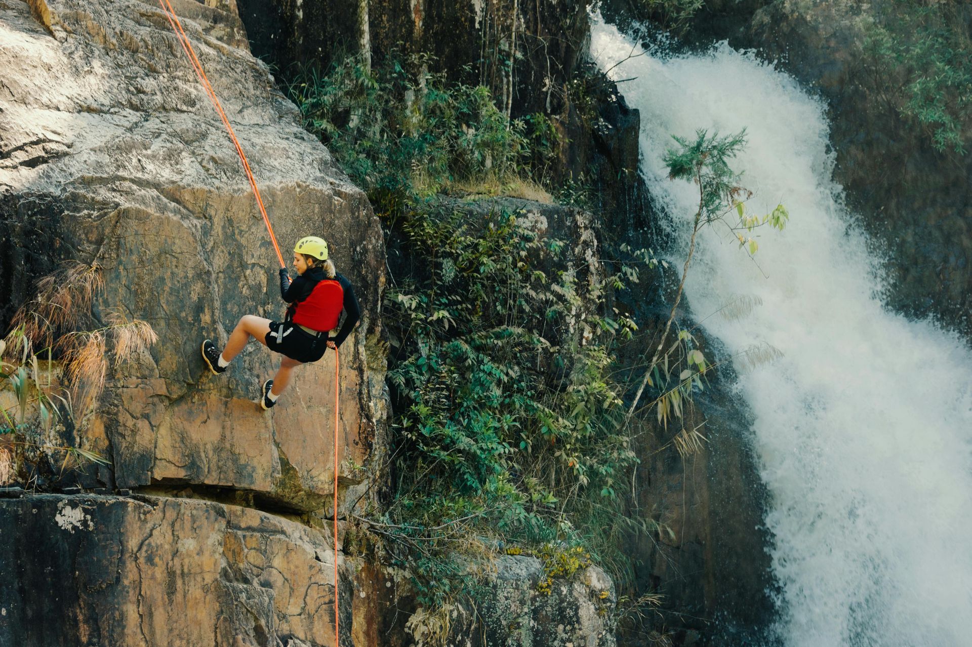 Person rappels down a rock face beside a rushing waterfall; they wear a helmet and harness.