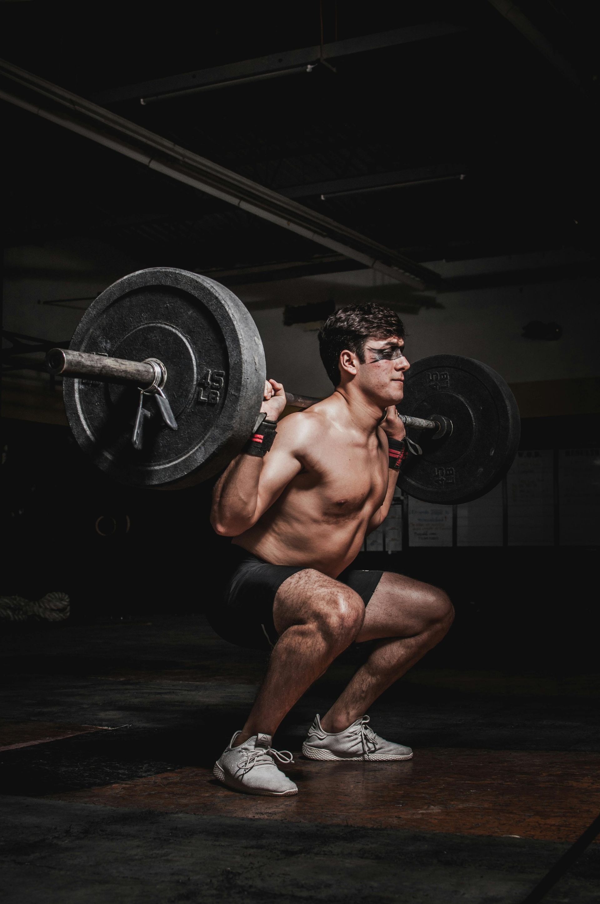 Man squats with a barbell in a dim gym. He is shirtless and wears black shorts and white shoes.
